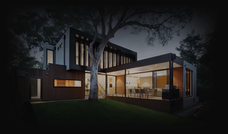 A modern home with warmly lit interiors beneath a large tree